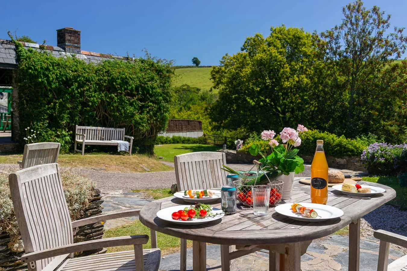 A rustic outdoor dining setup with wooden chairs and a round table, featuring fresh food, drinks, and flowers, set against a lush countryside backdrop.