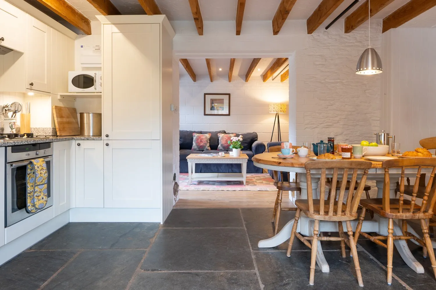 A cosy cottage kitchen and dining area with a wooden table set for breakfast, leading into a snug living space with a navy sofa, beamed ceiling, and soft lighting.