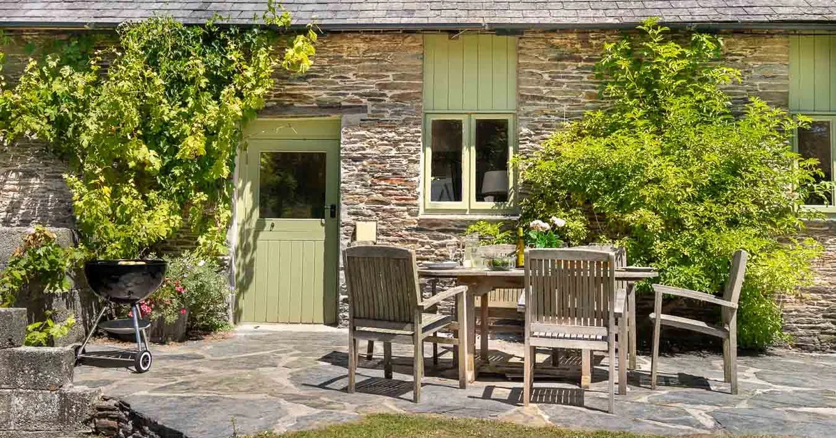 A charming stone cottage with green doors and windows, an outdoor dining area, and a barbecue, surrounded by lush greenery and bathed in sunlight.