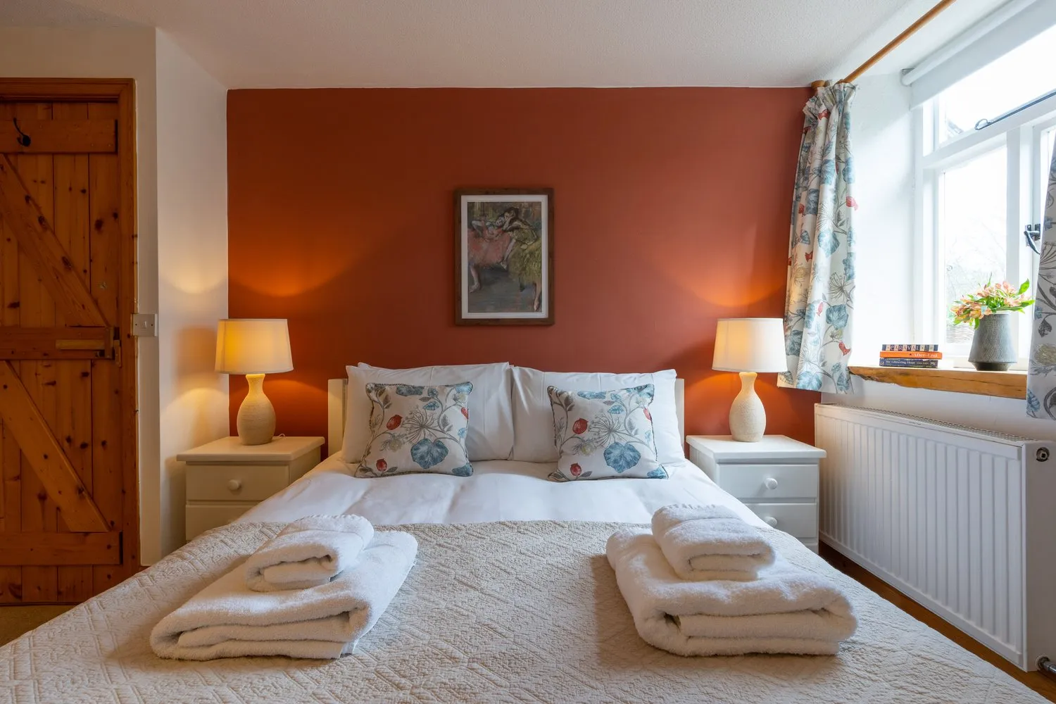 A cosy cottage bedroom with a double bed, warm lighting, a rustic wooden door, and a terracotta accent wall, creating a welcoming and relaxing atmosphere.