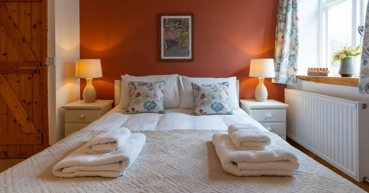 A cosy cottage bedroom with a double bed, warm lighting, a rustic wooden door, and a terracotta accent wall, creating a welcoming and relaxing atmosphere.