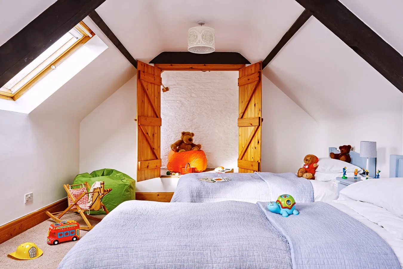 A cosy children's twin bedroom with soft blue bedding, wooden beams, skylights, and a play area featuring toys, a bean bag, and teddy bears for a welcoming feel.
