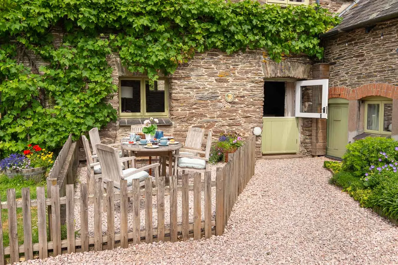 A charming stone cottage with a vine-covered wall, a quaint enclosed garden, and an outdoor dining area, featuring rustic wooden furniture and colourful flowers.