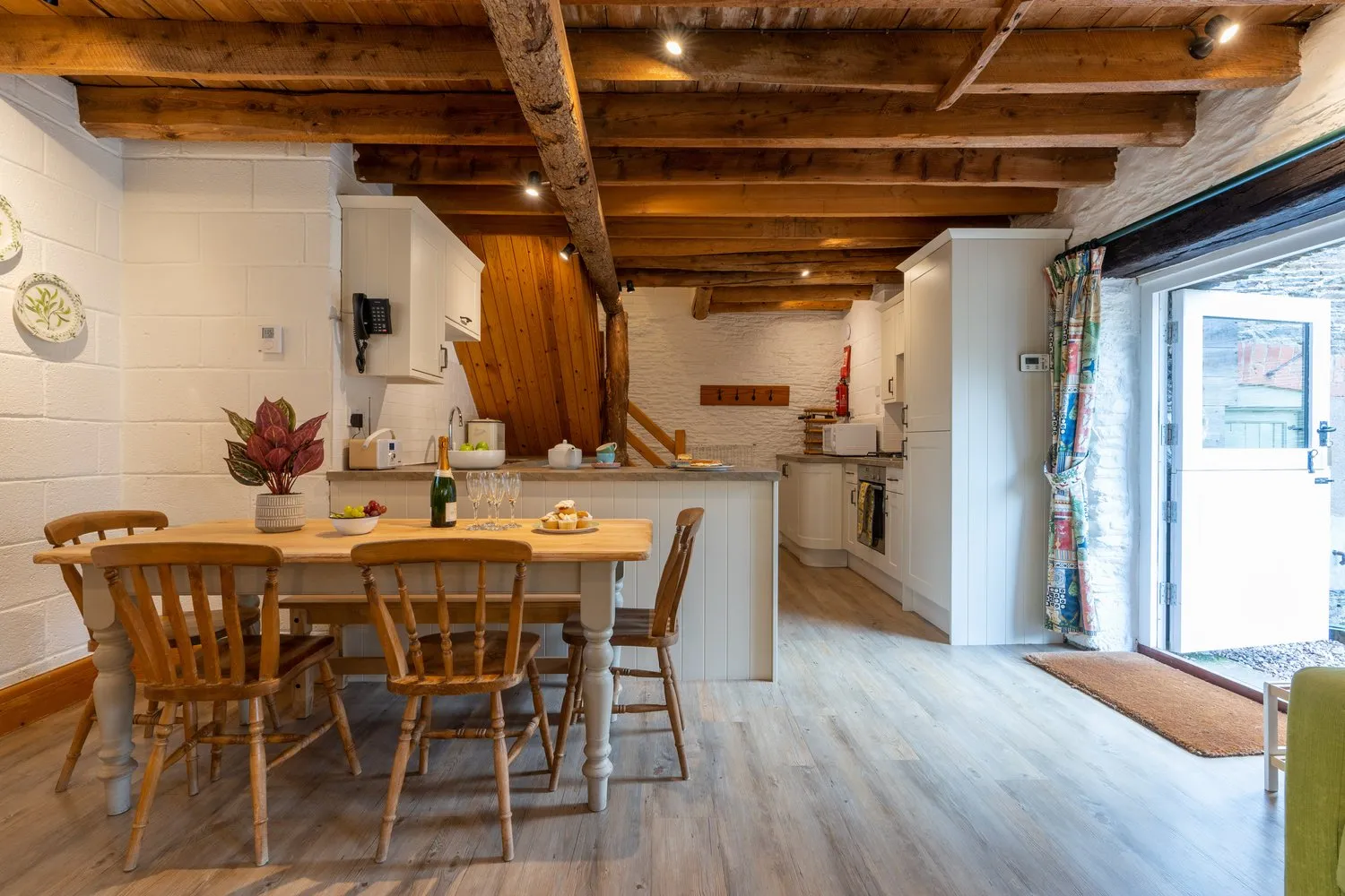 A rustic open-plan kitchen and dining area with exposed wooden beams, a farmhouse table, and a cosy atmosphere, leading to a bright outdoor space.