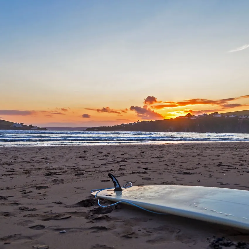 A surfboard rests on the sandy beach at sunset, with gentle ocean waves and a distant island in the background. The sky glows with warm orange and pink hues.