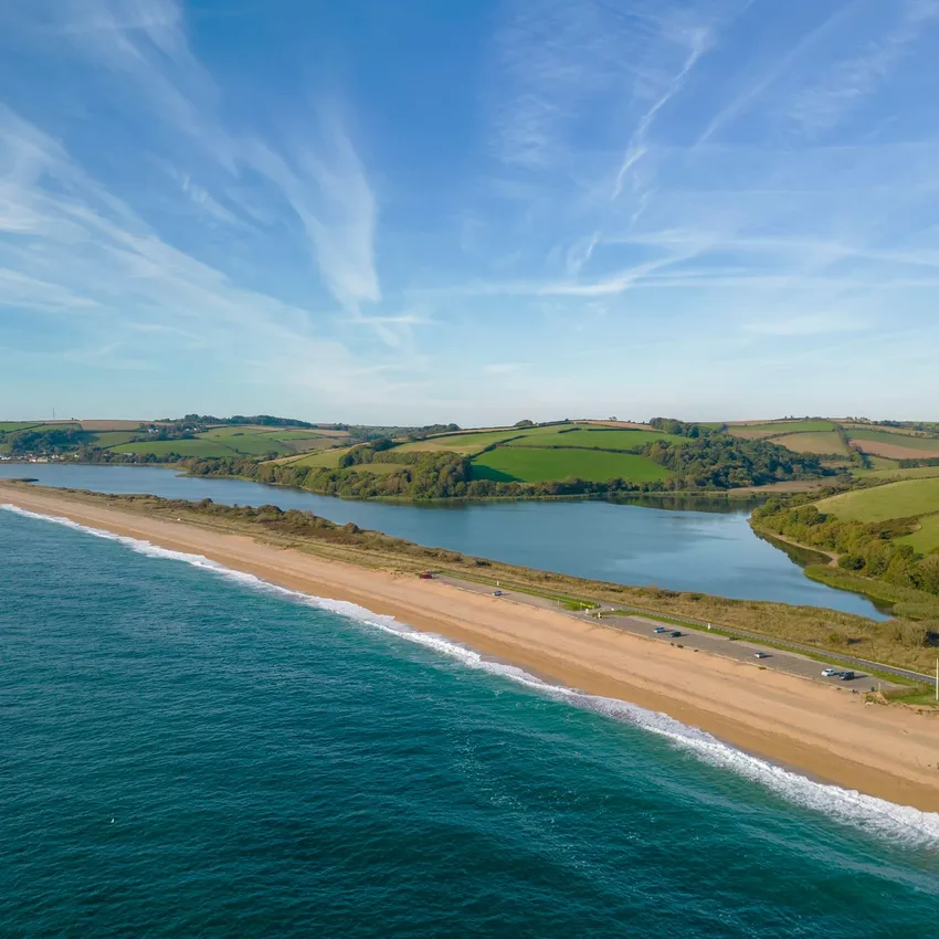 A coastal landscape with a long sandy beach, blue ocean waves, and a calm inland lagoon. Rolling green hills stretch under a clear blue sky with wispy clouds.