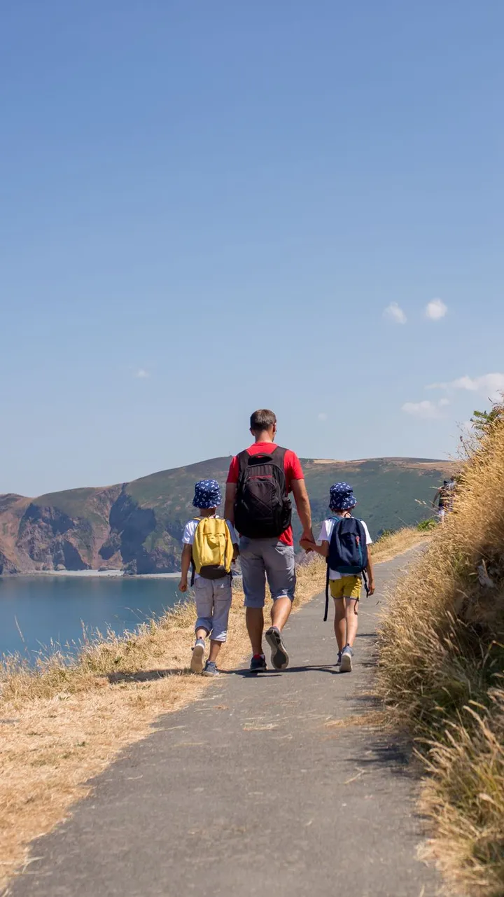 A man and two children, wearing backpacks, walk along a coastal path with a scenic view of cliffs and the ocean under a clear blue sky. Tall grass lines the trail.