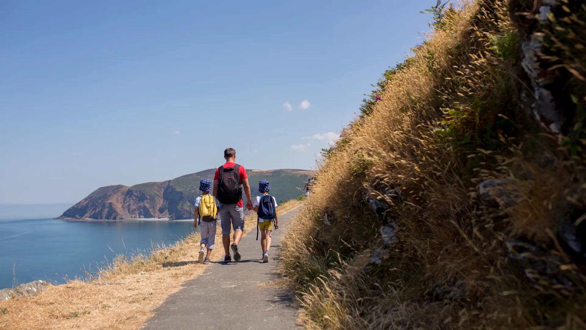 A man and two children, wearing backpacks, walk along a coastal path with a scenic view of cliffs and the ocean under a clear blue sky. Tall grass lines the trail.