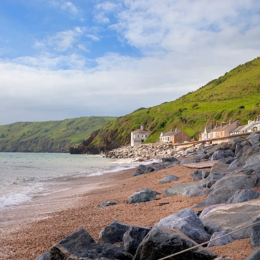 A coastal village with pastel-colored houses sits at the base of lush green hills, overlooking a rocky beach where gentle waves lap against the shore under a partly cloudy sky.