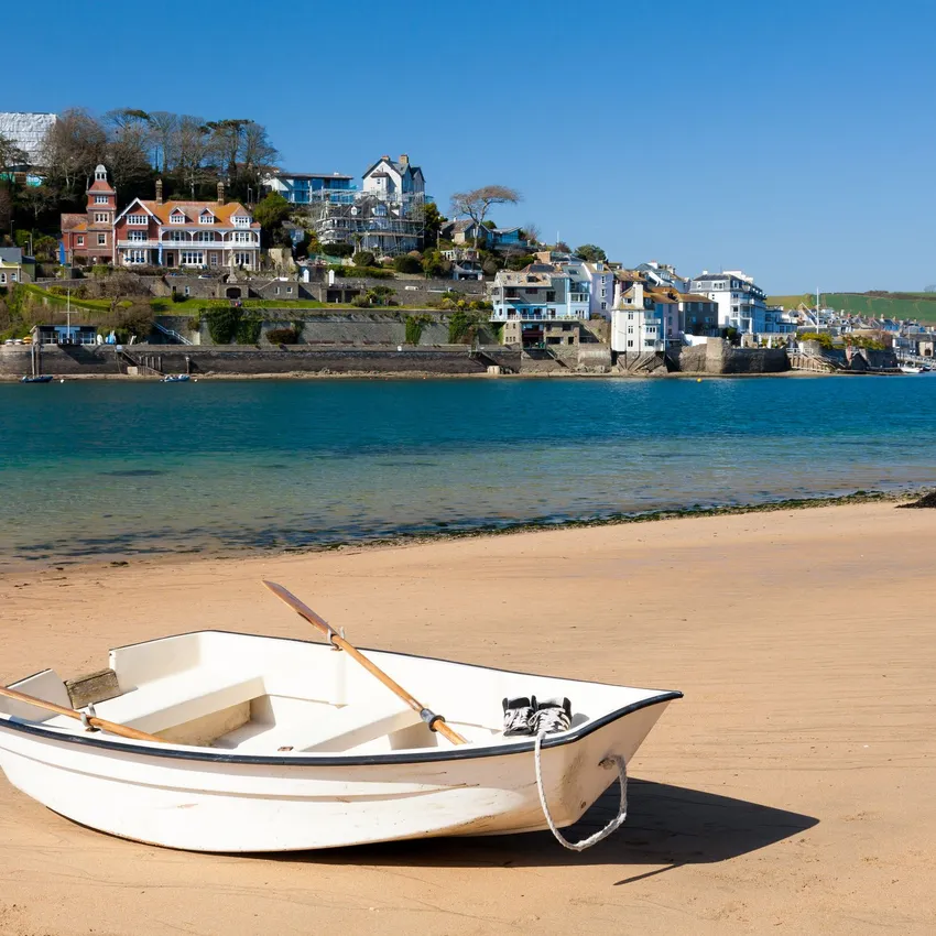 A white rowboat with wooden oars rests on a sandy beach, with calm blue waters and a hillside town of colorful houses and greenery in the background.