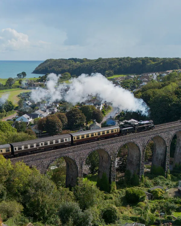 A steam train crosses a stone viaduct, releasing white smoke into the sky. Below, a village with green fields stretches toward the blue sea in the background.