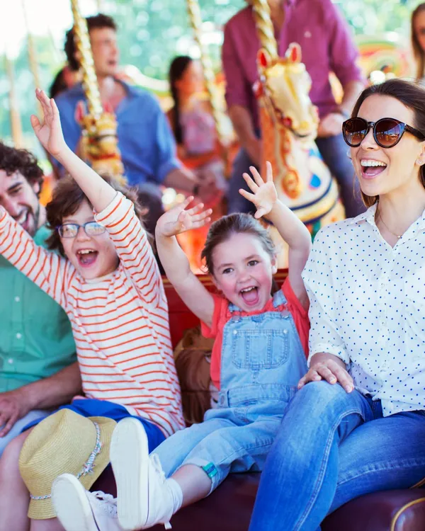 A joyful family rides a vintage carousel, with children raising their hands in excitement. The brightly colored horses and ornate decorations add to the festive atmosphere.