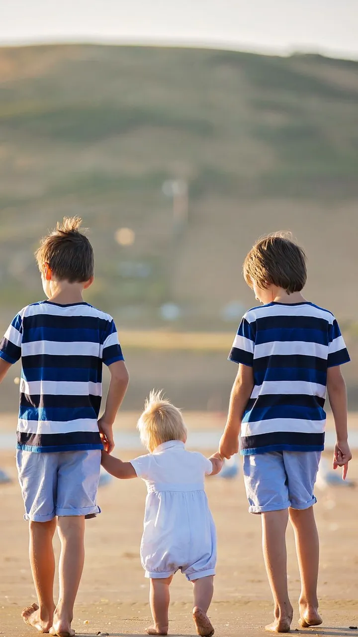 Three young siblings walk barefoot on a sandy beach, with the older two holding the toddler’s hands. The golden light and rolling hills create a peaceful backdrop.