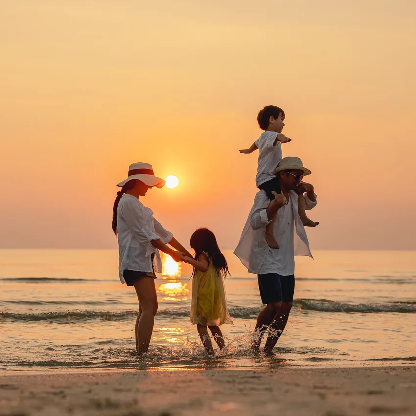 A family enjoys a sunset walk along the beach, splashing in the shallow waves. The parents wear white shirts, with one child on a father's shoulders and another playing.