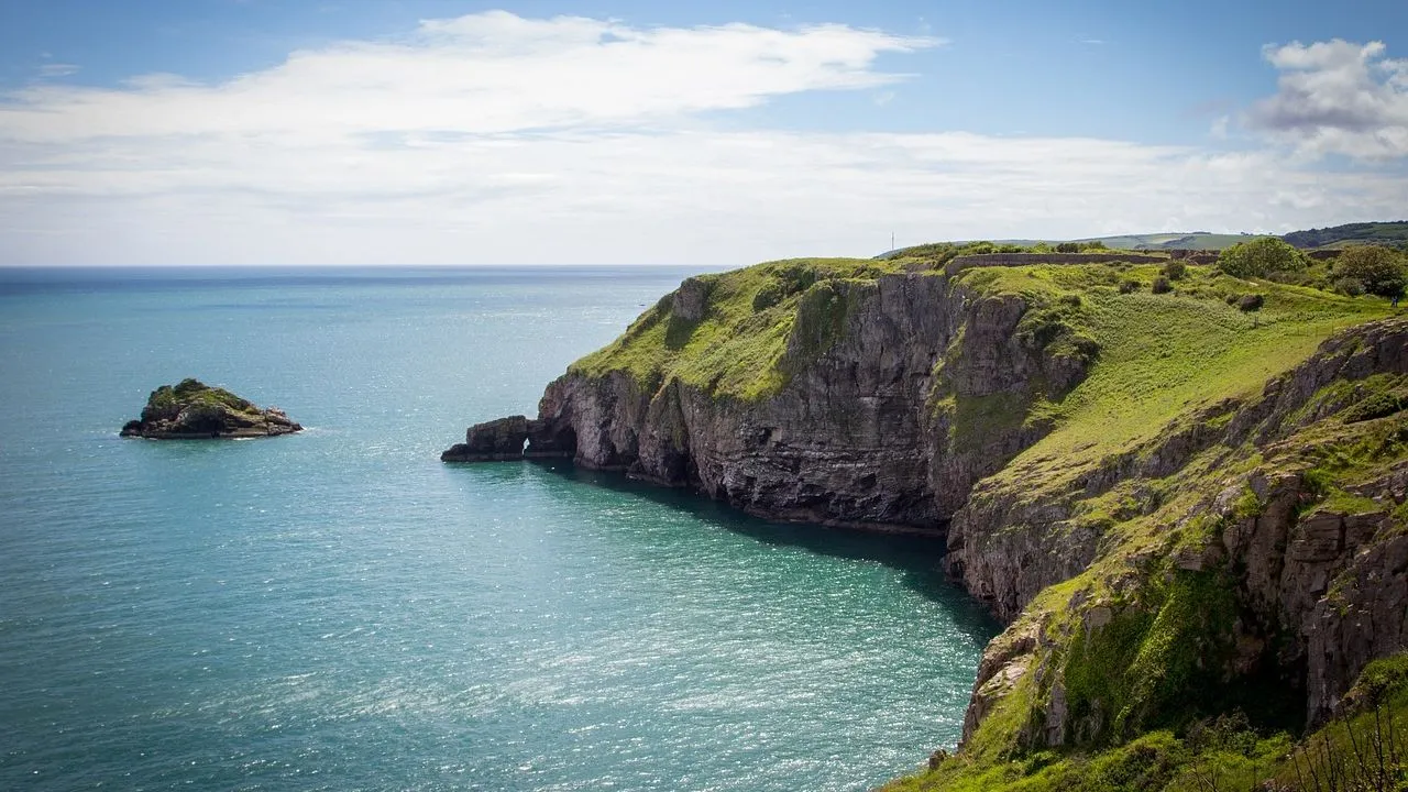 Rocky cliffs covered in green grass overlooking a calm turquoise sea, with a small island nearby under a partly cloudy blue sky.