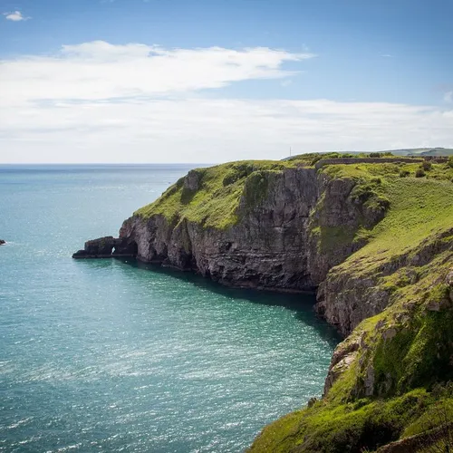 Rocky cliffs covered in green grass overlooking a calm turquoise sea, with a small island nearby under a partly cloudy blue sky.