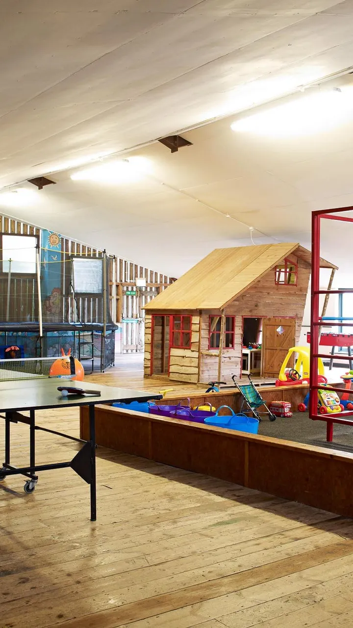Indoor play area with a wooden playhouse, climbing frame, trampoline, ping pong table, and various toys on a wooden floor under a sloped ceiling.