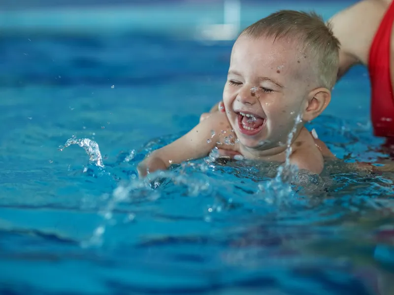 A joyful baby splashes in a swimming pool, laughing with eyes closed, while being supported by an adult wearing a red swimsuit in the background.