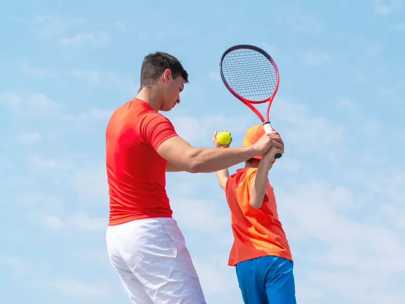 A tennis coach in a red shirt guides a young child in an orange outfit on how to swing a racket, against a bright blue sky with scattered clouds.