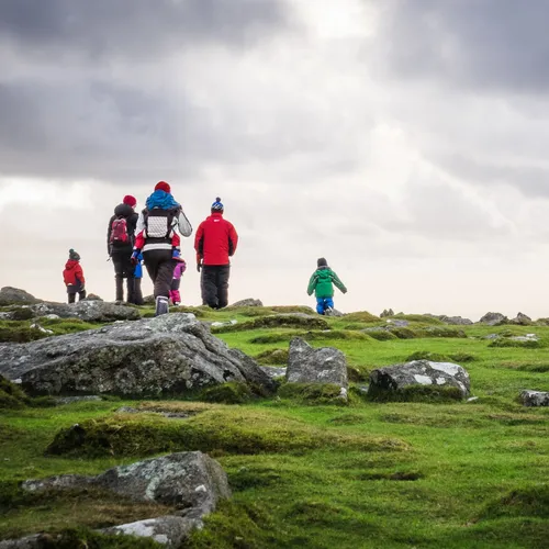 A family, including children, hike across a rocky, grassy moorland under a cloudy sky, dressed in bright winter gear.
