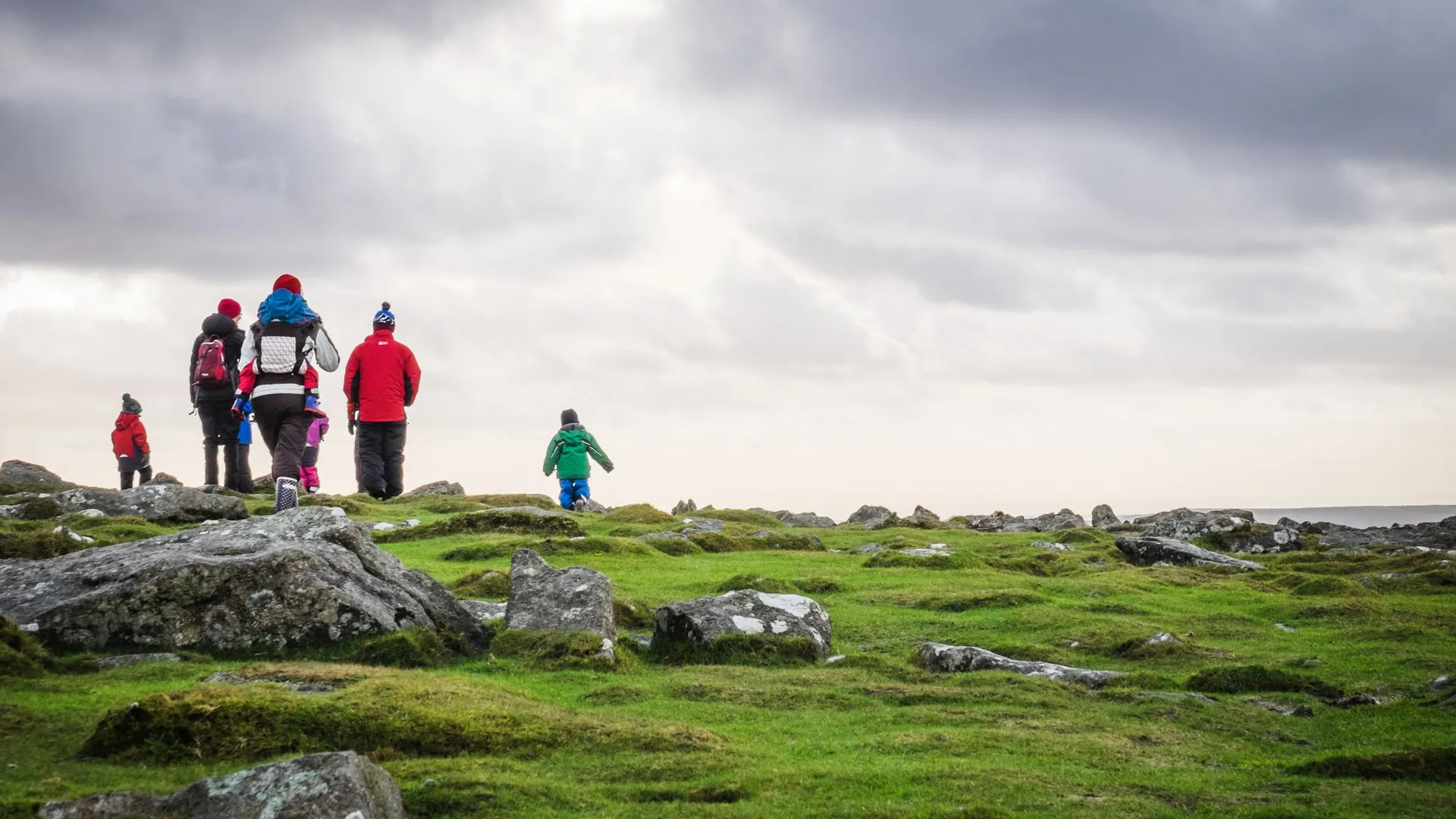 A family, including children, hike across a rocky, grassy moorland under a cloudy sky, dressed in bright winter gear.