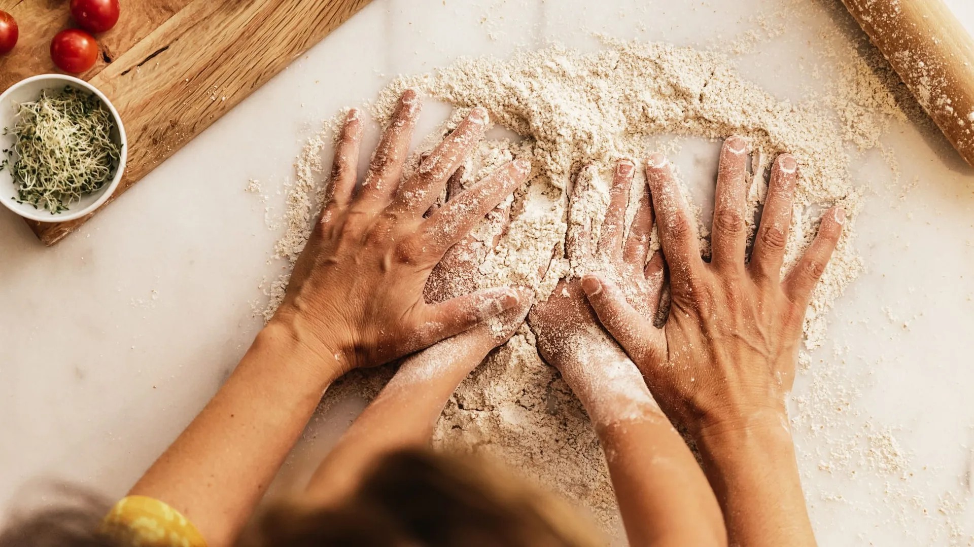 Adult and child hands working together to knead flour on a counter, surrounded by fresh ingredients like tomatoes, herbs, and a rolling pin.