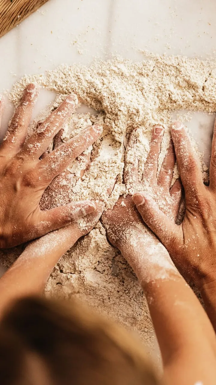 Adult and child hands working together to knead flour on a counter, surrounded by fresh ingredients like tomatoes, herbs, and a rolling pin.