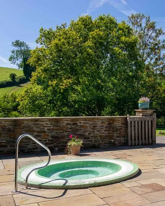A serene outdoor spa area with a round hot tub, two wicker loungers, and a stone wall, surrounded by lush greenery and rolling hills under a bright blue sky.