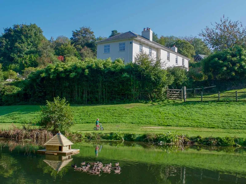 A scenic countryside view with a pond, a floating duck house, and a group of ducks. A child rides a bike on a grassy path, with a white house nestled among trees.