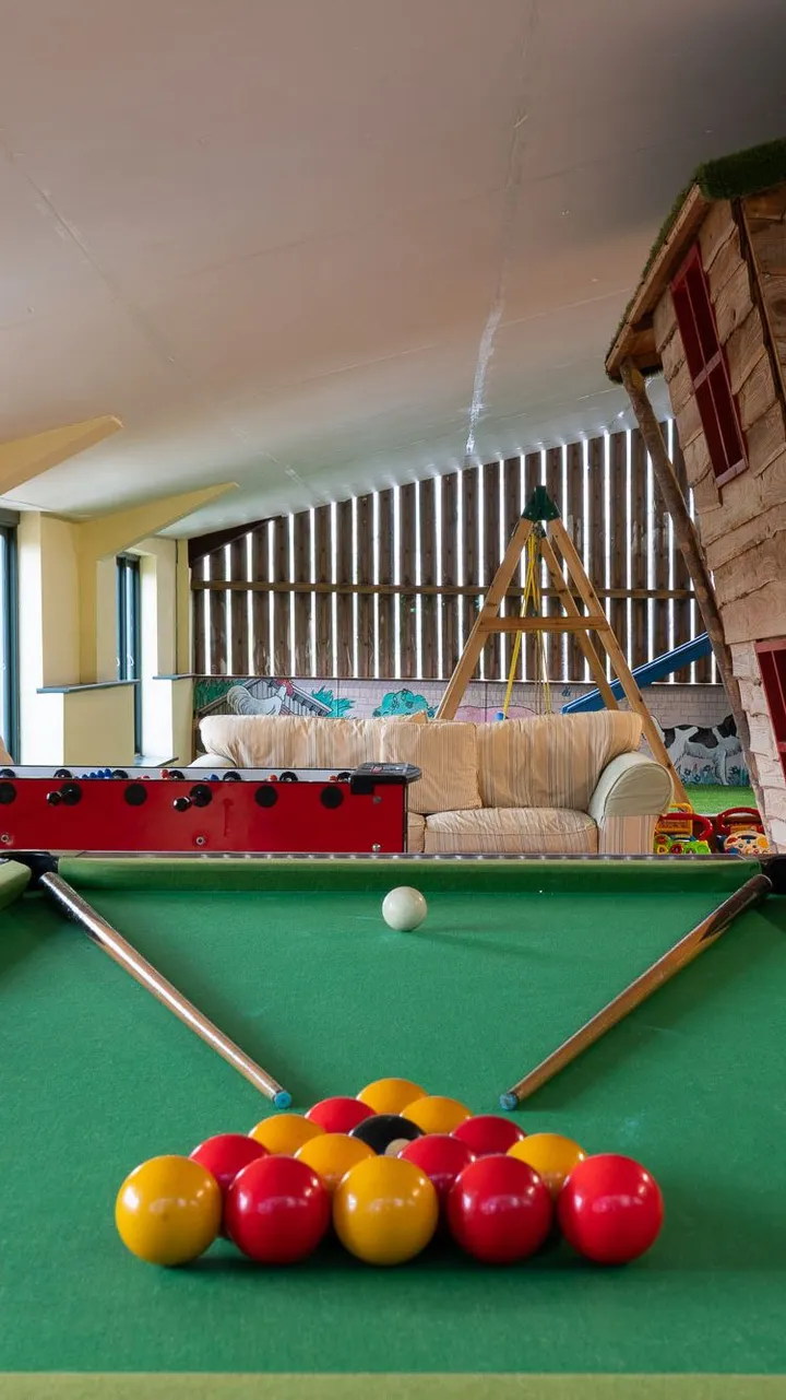 A games room with a green-felt pool table in the foreground, a foosball table, sofas, a wooden playhouse, and a climbing frame under a sloped ceiling with large windows.