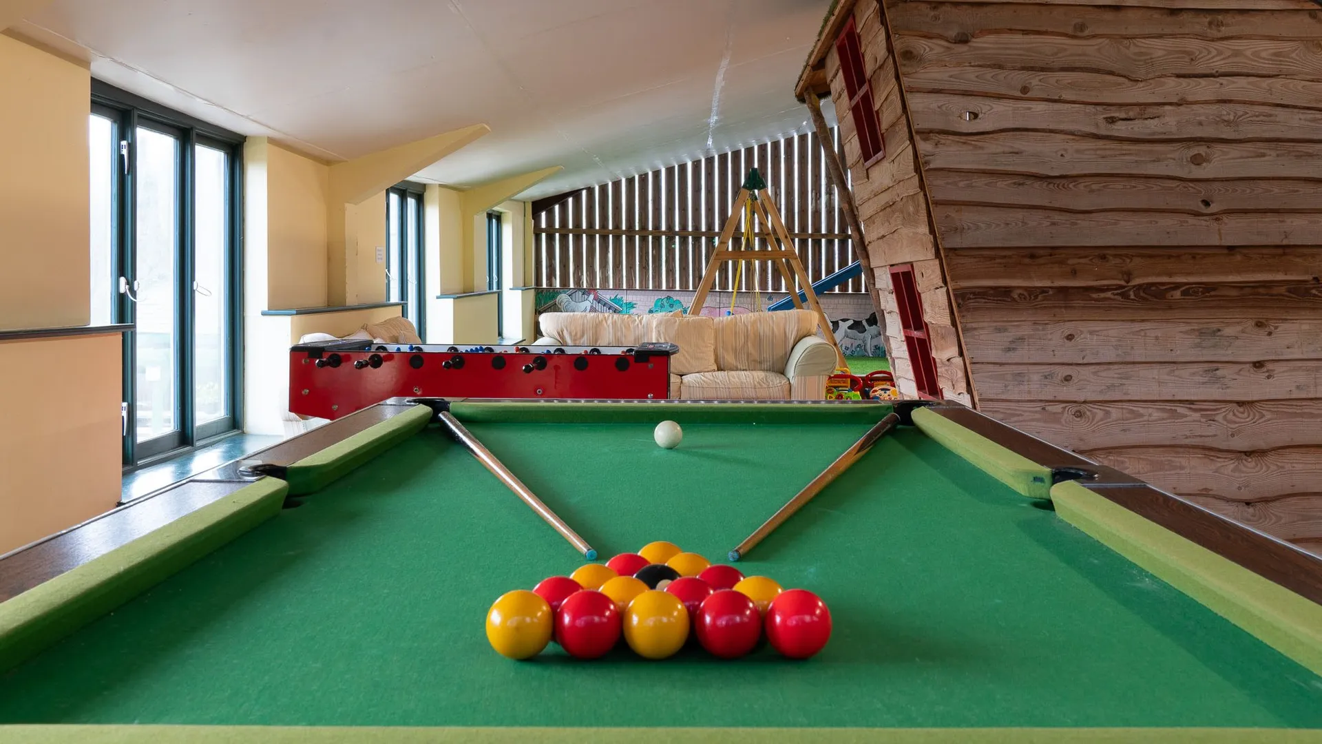 A games room with a green-felt pool table in the foreground, a foosball table, sofas, a wooden playhouse, and a climbing frame under a sloped ceiling with large windows.