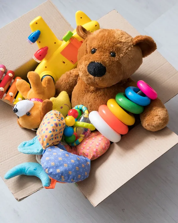 A cardboard box filled with toys, including a teddy bear, stacking rings, and a toy airplane, sits on a light wooden floor with more plush toys and vehicles scattered nearby.