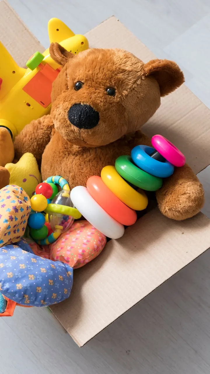 A cardboard box filled with toys, including a teddy bear, stacking rings, and a toy airplane, sits on a light wooden floor with more plush toys and vehicles scattered nearby.