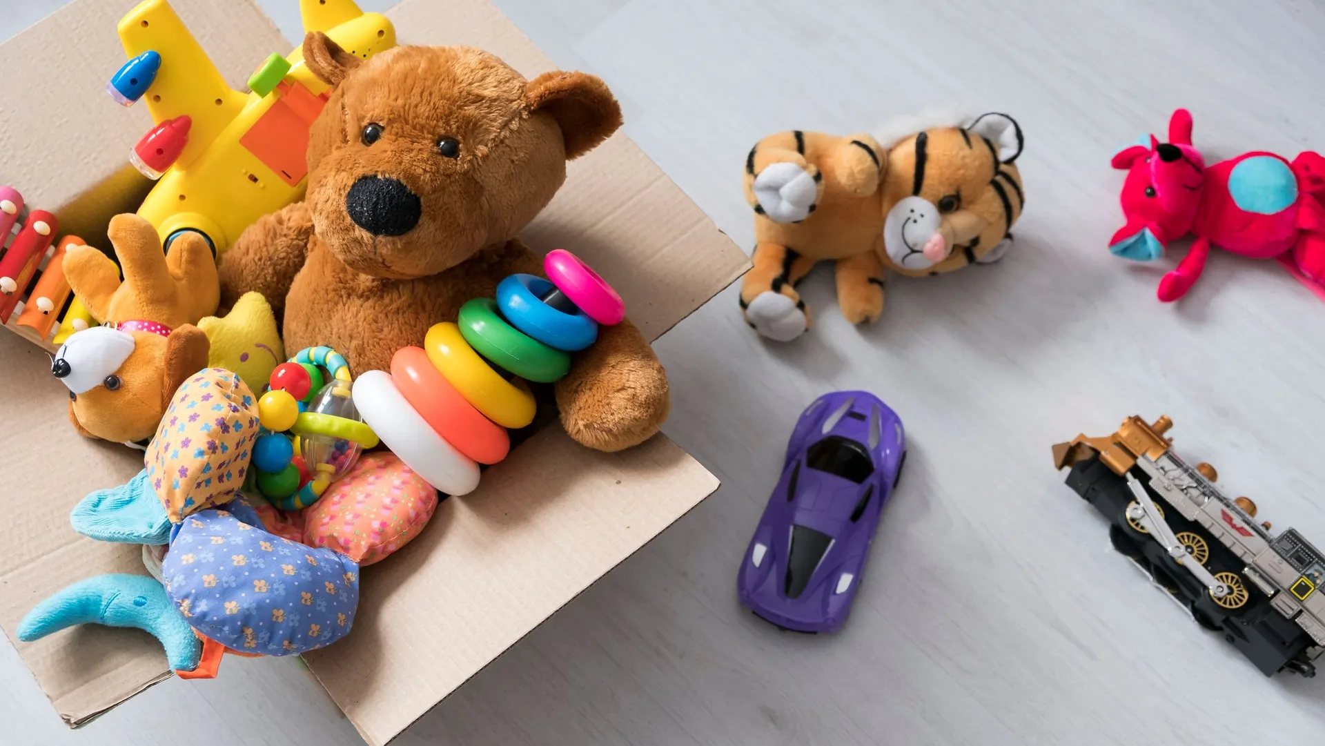 A cardboard box filled with toys, including a teddy bear, stacking rings, and a toy airplane, sits on a light wooden floor with more plush toys and vehicles scattered nearby.