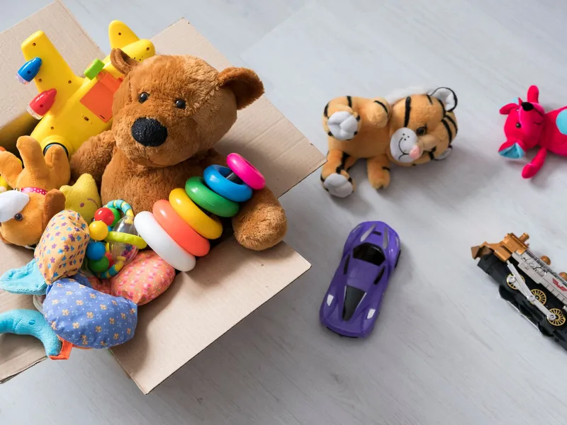 A cardboard box filled with toys, including a teddy bear, stacking rings, and a toy airplane, sits on a light wooden floor with more plush toys and vehicles scattered nearby.