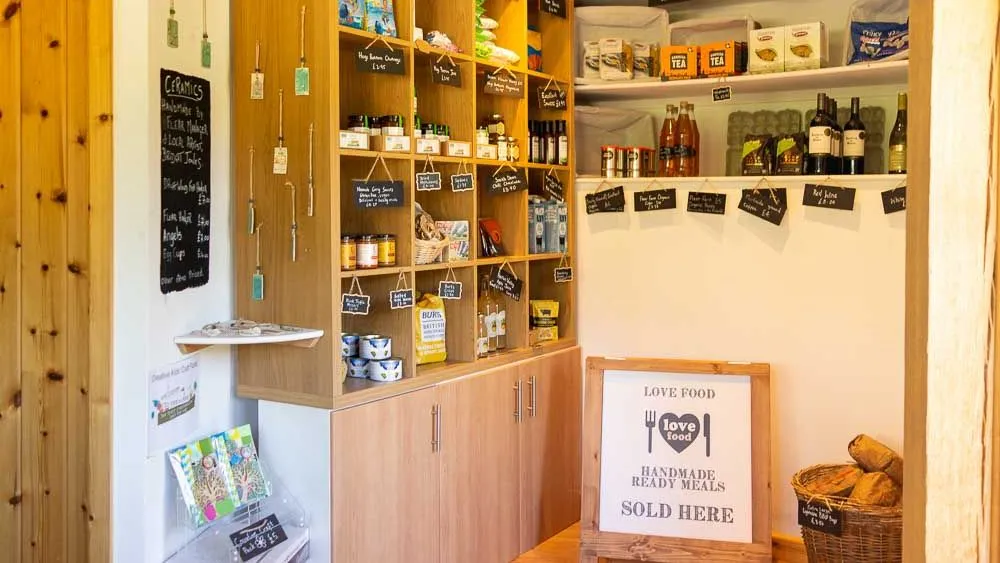 A small farm shop with wooden shelves stocked with local goods, including jams, tea, pasta, and wine, with handwritten labels and a sign for handmade ready meals.