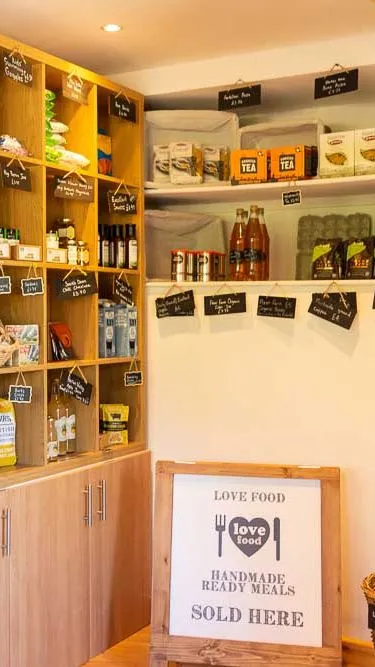 A small farm shop with wooden shelves stocked with local goods, including jams, tea, pasta, and wine, with handwritten labels and a sign for handmade ready meals.