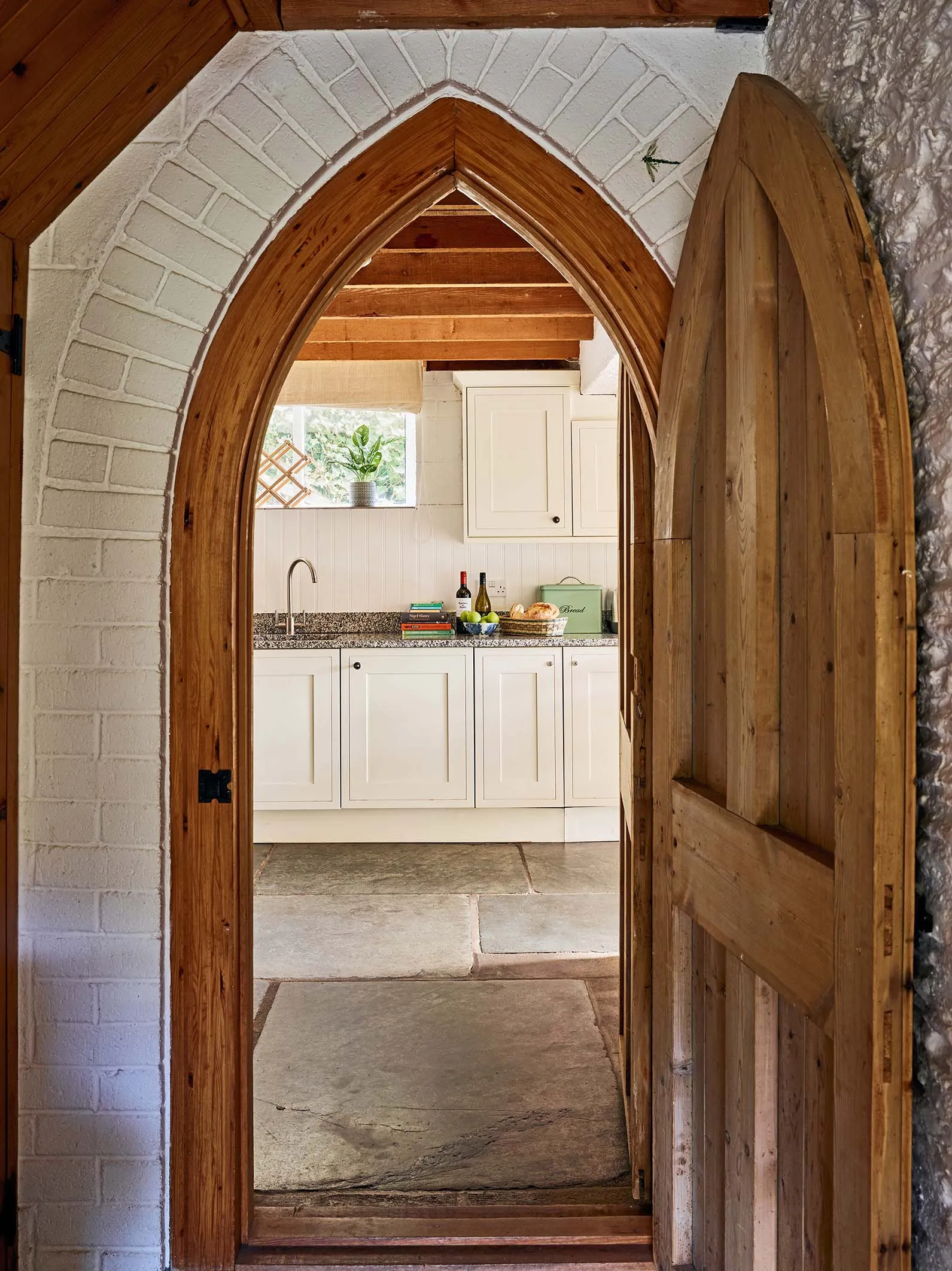 View into the kitchen at Duck’s Quarry, Flear Farm Cottages, through a pointed arched wooden door, with stone flooring and cream cabinetry beyond.