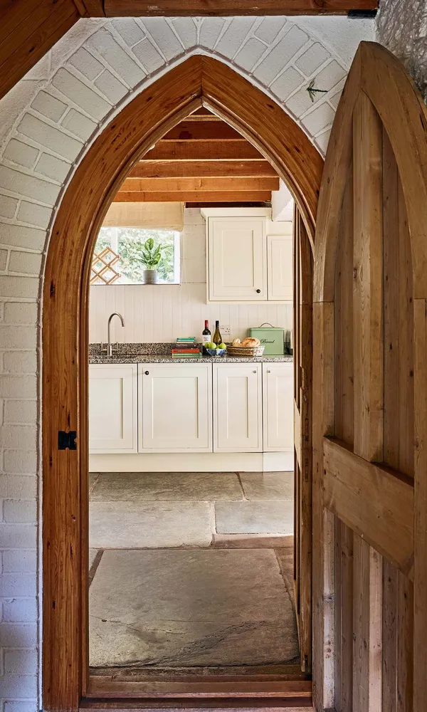View into the kitchen at Duck’s Quarry, Flear Farm Cottages, through a pointed arched wooden door, with stone flooring and cream cabinetry beyond.