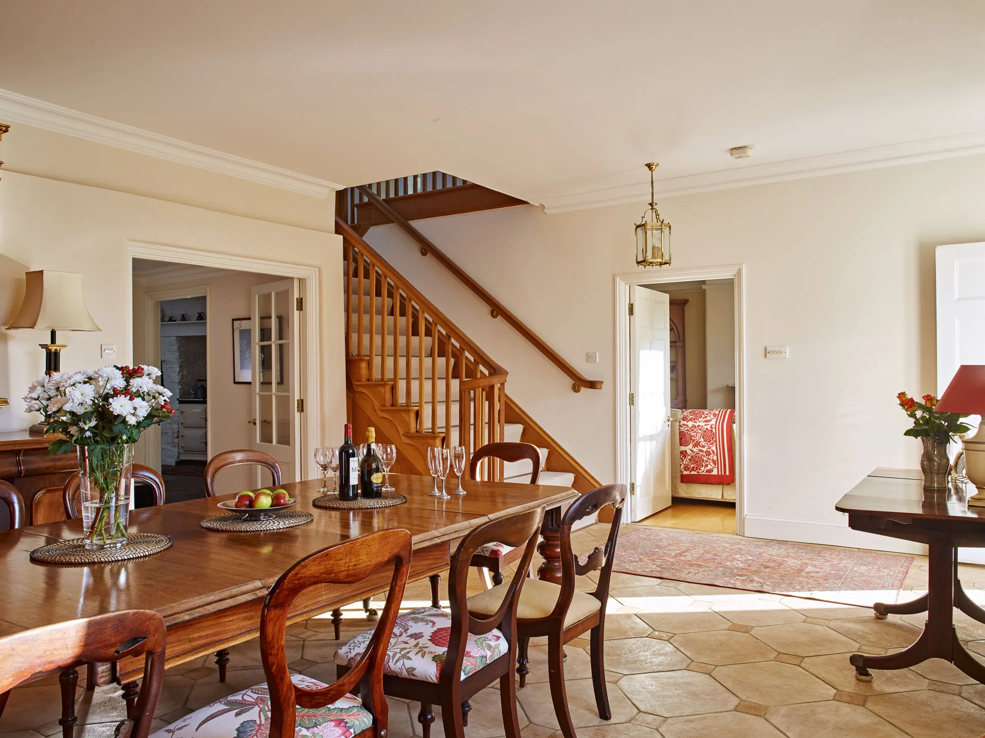 A warm, elegant dining room with a wooden table set with wine, glasses, and flowers, featuring classic chairs, a staircase, and an open doorway leading to a bedroom.