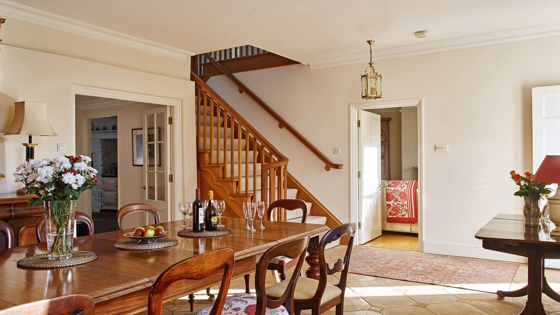 A warm, elegant dining room with a wooden table set with wine, glasses, and flowers, featuring classic chairs, a staircase, and an open doorway leading to a bedroom.