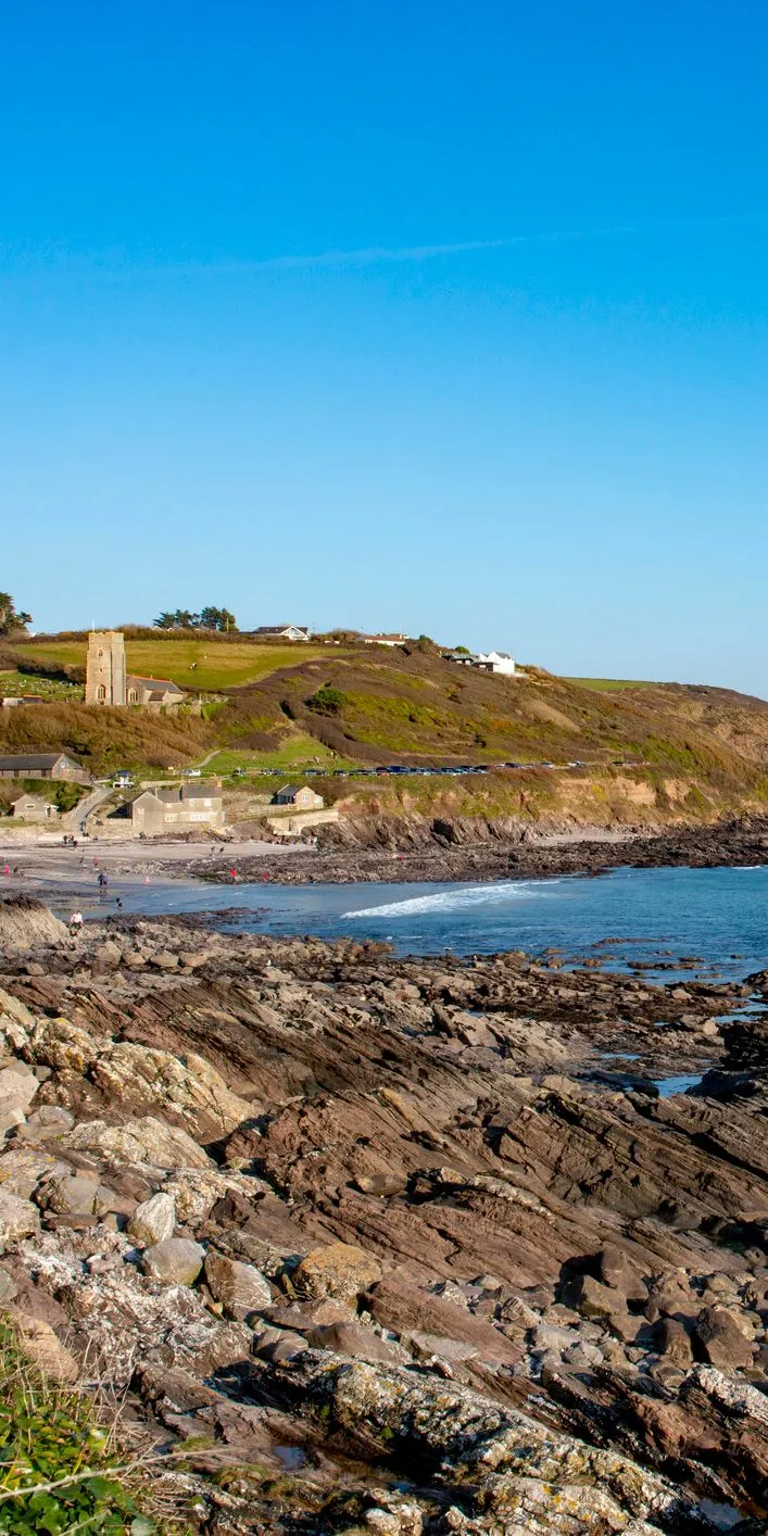 Rocky shoreline at Wembury Beach in Wembury, Devon, with calm blue sea, distant cliffs, and a church tower overlooking the coastal landscape.