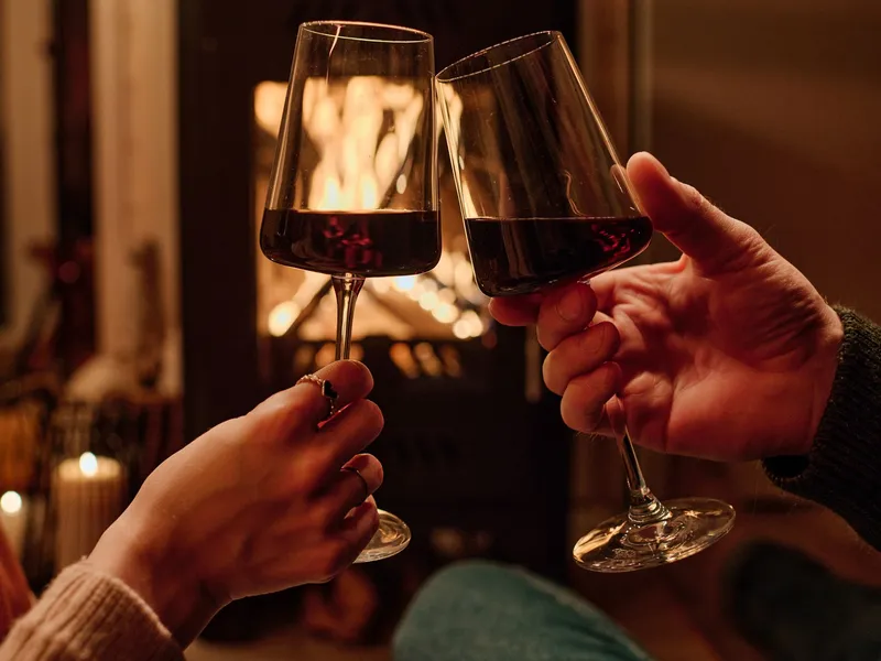 Two people clinking glasses of red wine in front of a cozy fireplace.