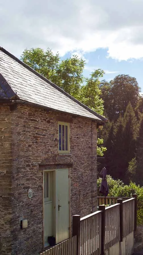 A rustic stone cottage with a slate roof sits beside a wooden fence, overlooking a lush green valley filled with dense trees under a partly cloudy sky.