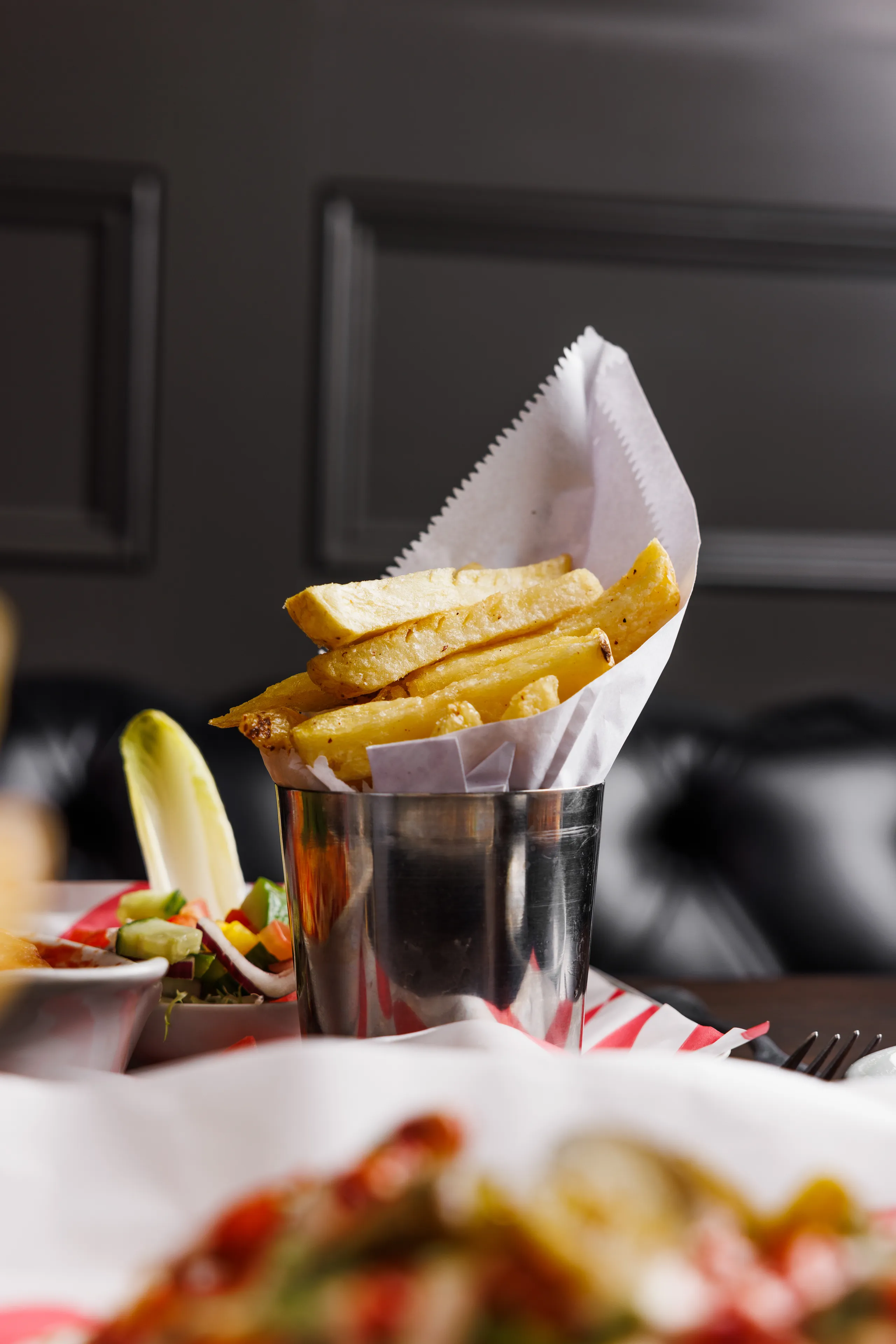 Golden thick-cut chips served in a metal cup lined with white paper beside a fresh salad.