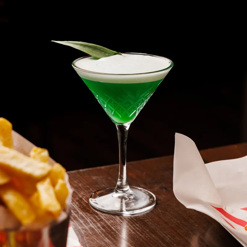 Bright green cocktail with a creamy white foam top and leaf garnish in a patterned stemmed glass