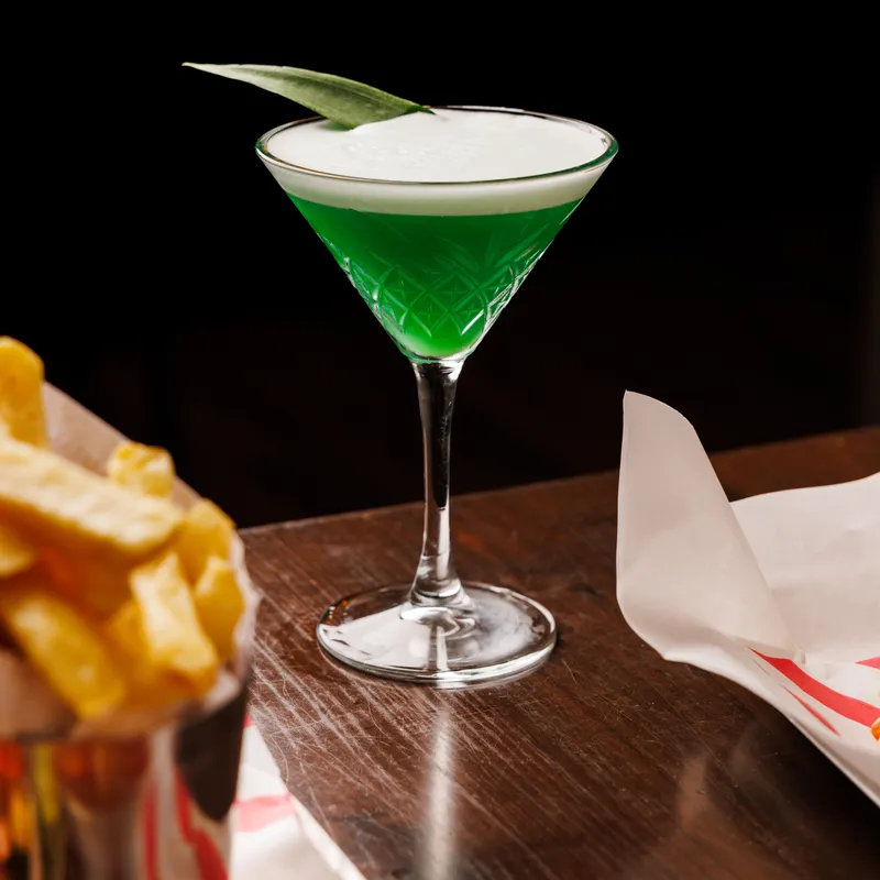 Bright green cocktail with a creamy white foam top and leaf garnish in a patterned stemmed glass