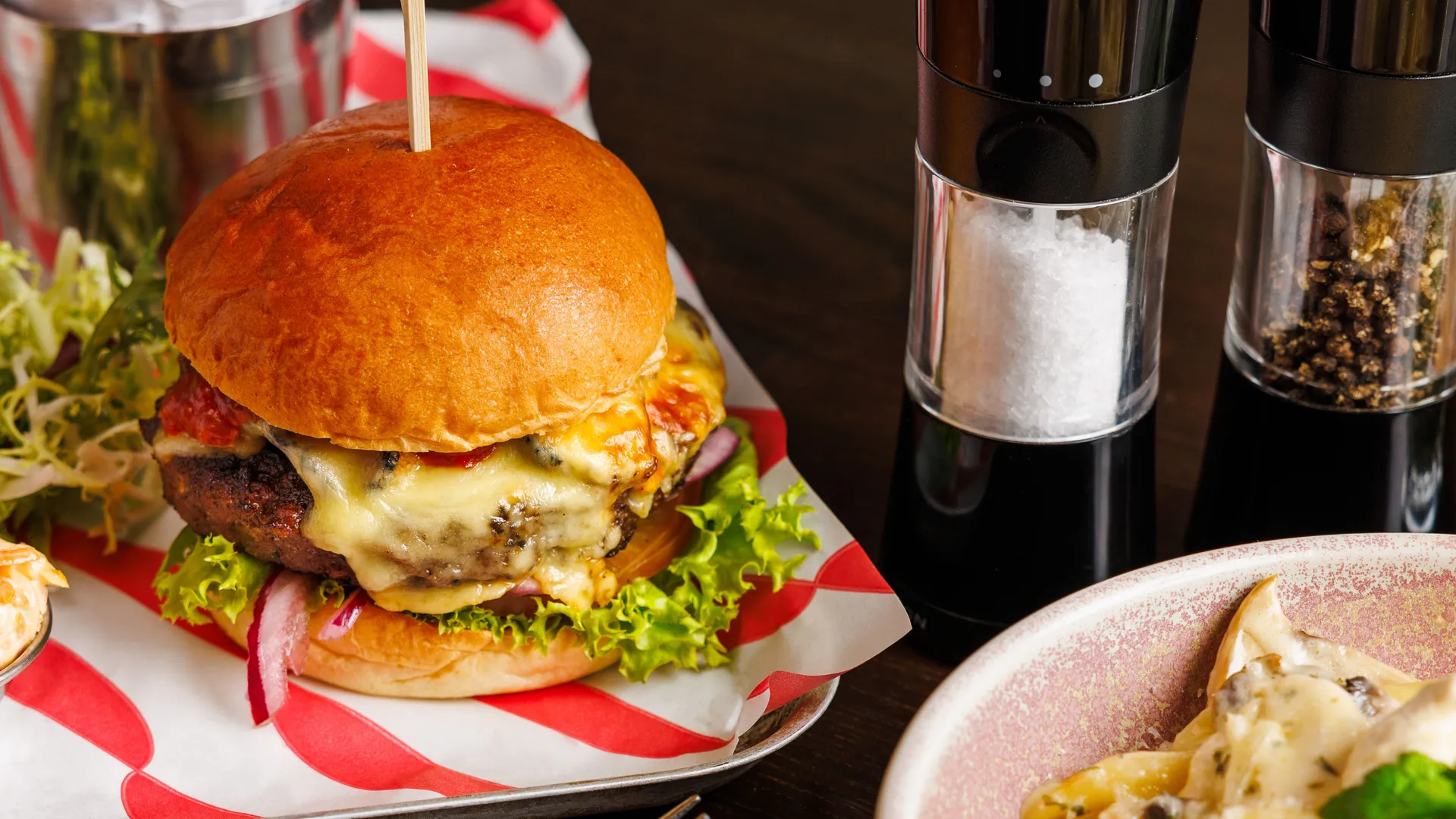 Cheeseburger with lettuce and red onion served with chips, salt and pepper grinders, and a bowl of creamy pasta.