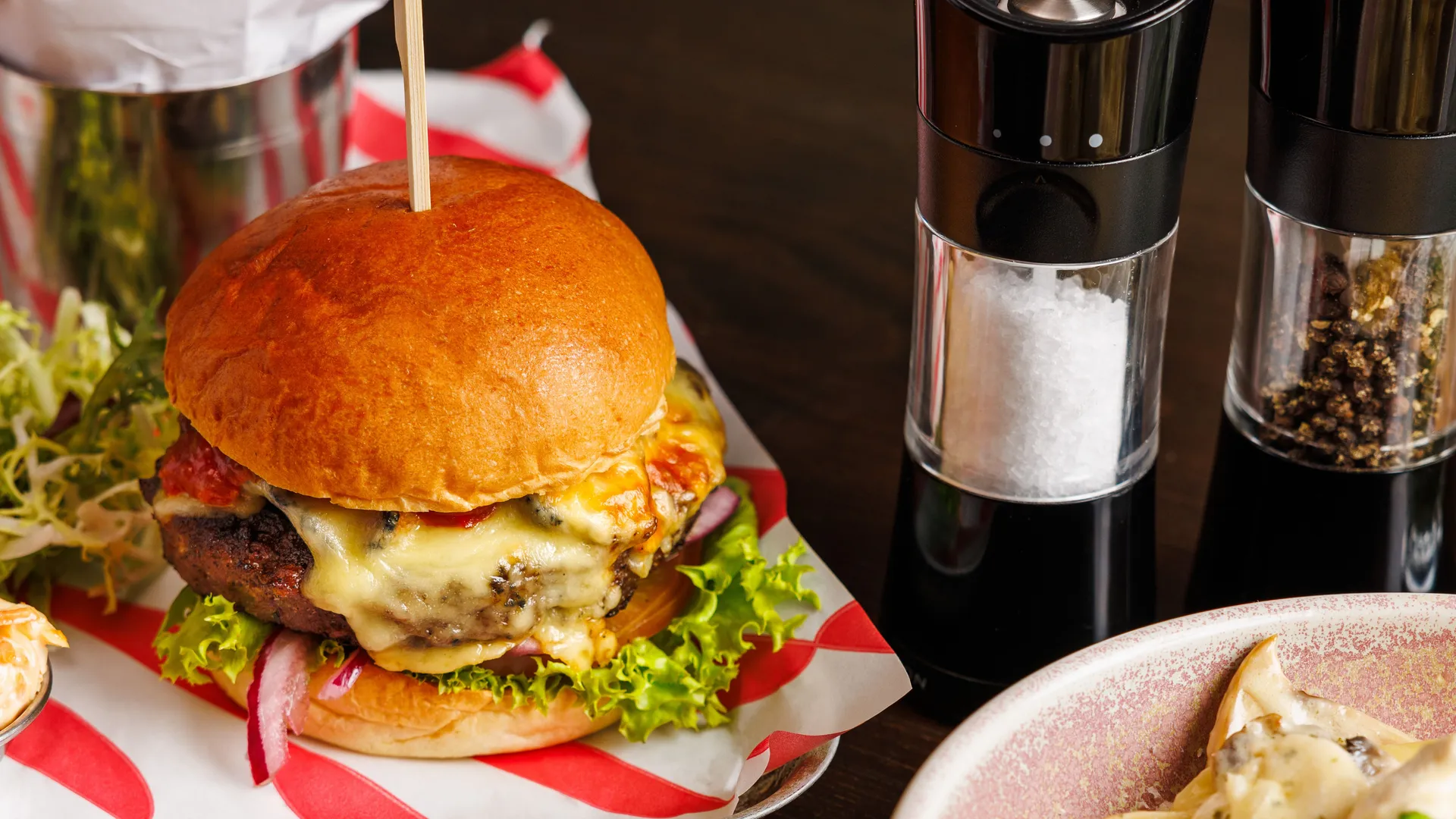 Cheeseburger with lettuce and red onion served with chips, salt and pepper grinders, and a bowl of creamy pasta.