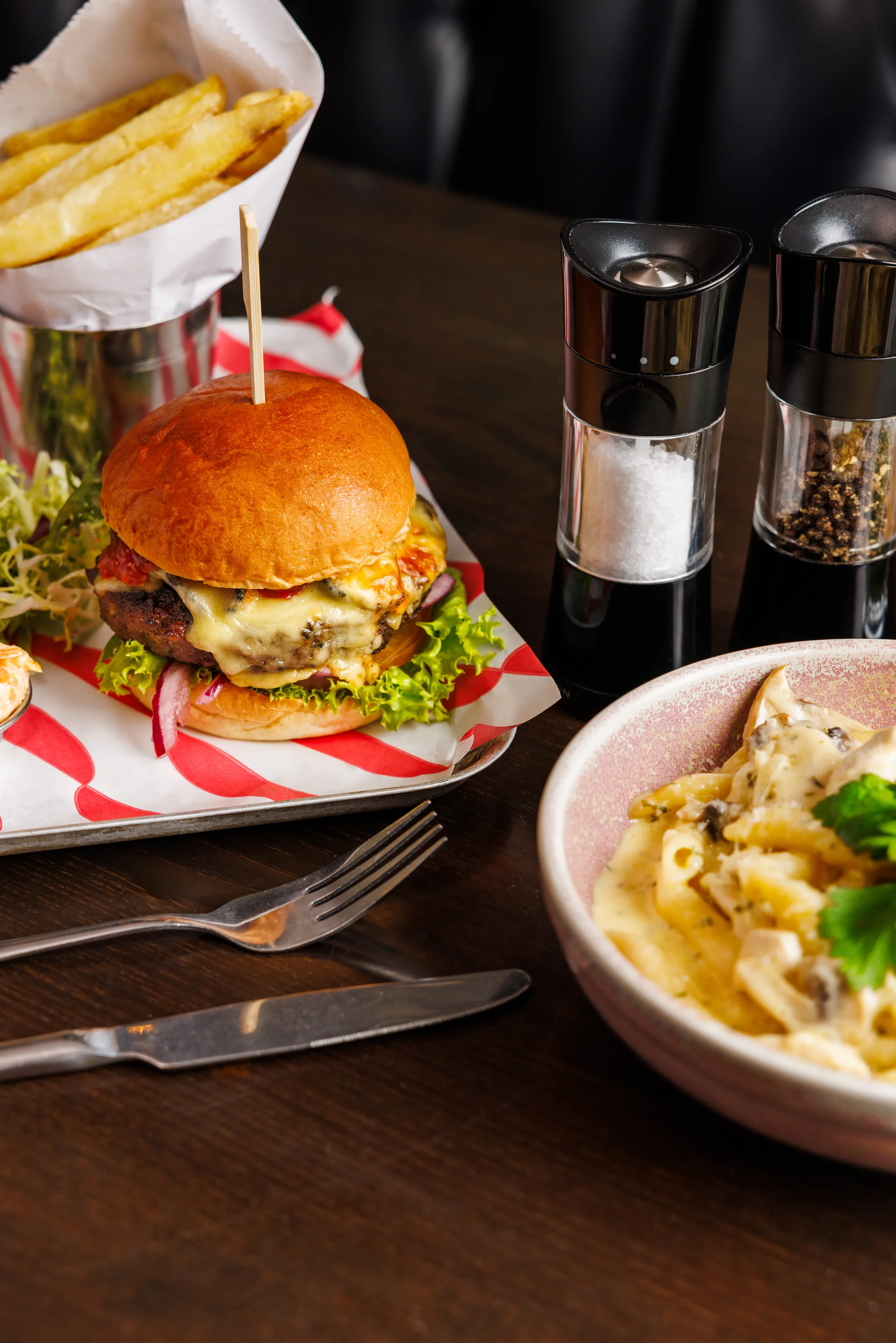 Cheeseburger with lettuce and red onion served with chips, salt and pepper grinders, and a bowl of creamy pasta.